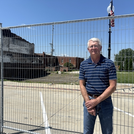 Man with gray hair wearing a blue polo with white stripes standing in front of a fence with an empty lot in the background