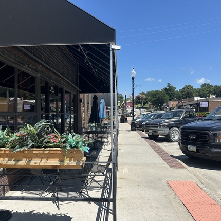 A downtown street with a building on the left with table and chairs in front and cars parked on a street on the right