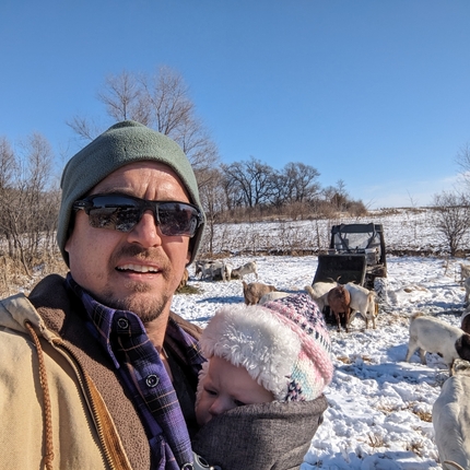Hombre con un abrigo marrón y una gorra verde sosteniendo a un bebé envuelto en una manta oscura con un sombrero rosa y blanco. Detrás de él hay un campo cubierto de nieve con cabras pastando en el heno que él colocó.