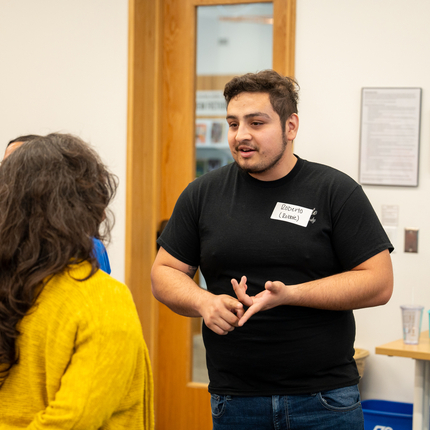 Hi dark haired male wearing a black short sleeve shirt and blue jeans talks to someone with long brown hair, wearing a yellow sweater 