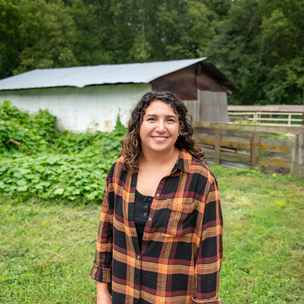 Woman with dark hair smiling at the camera, wearing an orange and black plaid long sleeve shirt. Background is an outbuilding with a wooden fence in front and a vine plant on the side.