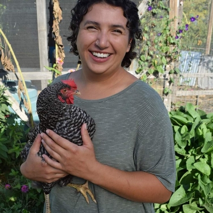 Woman with dark hair, smiling at the camera, wearing a short sleeved gray shirt, holding a chicken. An outbuilding in the background with plants.