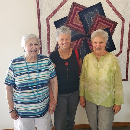 Three older women standing in front of a hand-made quilt.