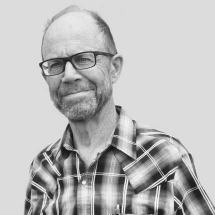 Man with balding hair, black large-rimmed glasses, wearing a dark plaid shirt, smiling at camera