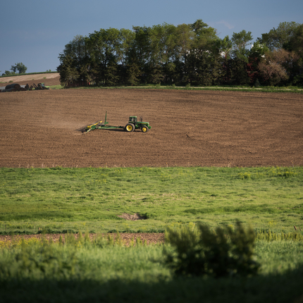 Un tractor tirant d'una jardinera, plantant en un camp nu de terra marró. Amb una zona d'herba en primer pla i una línia d'arbres a l'altre costat del camp.