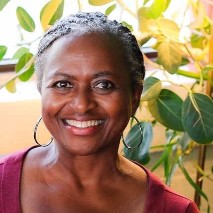 Mujer negra sonriendo directamente a la cámara. Lleva el pelo recogido hacia atrás, lleva unos grandes pendientes de aro, lleva una camiseta de color fucsia y hay plantas de fondo.