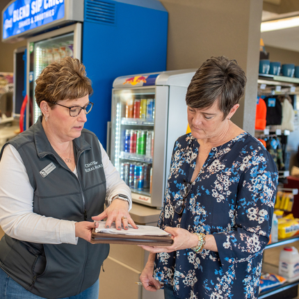 Dos mujeres blancas con pelo corto color café están dentro de una tienda de conveniencia paradas en frente de una maquina grande color azul y una nevera para bebidas a sus espaldas mientras miran hacia unos papeles en mano.
