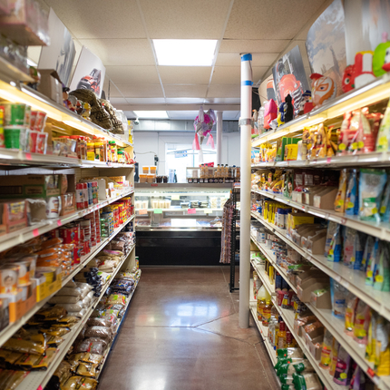 Aisle at a locally owned grocery store with shelves of food products on both sides of the aisle, and a meat counter at the front