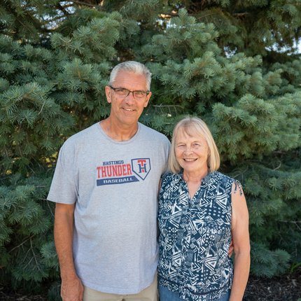 A man and woman in front of a tree, looking at the camera. The man has short white hair, glasses, and a gray T-shirt. The woman has shoudler-length blonde hair and is wearing a blue and white tank top.