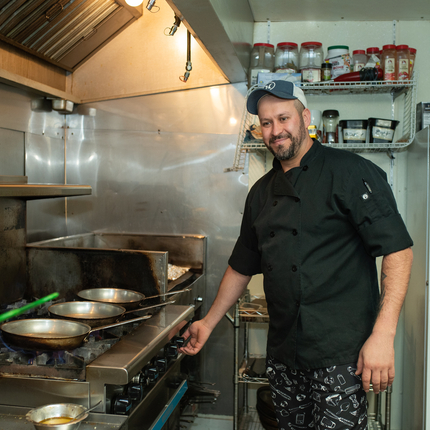 Un hombre esta parado en seguida de una estufa con tres sartenes en los quemadores. El hombre esta portando una cachucha de béisbol, una camisa de chef color negro. Hay especies a su espalda y un refrigerador color blanco.