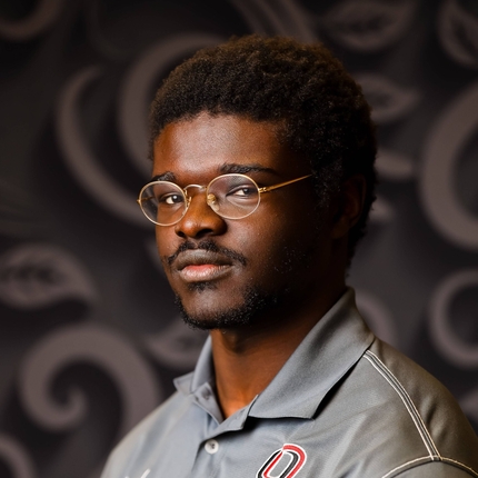 Peter David, un jove afroamericà que porta ulleres rodones amb marc daurat, posa per una imatge davant d'un fons fosc amb un polo gris amb un logotip vermell i negre al costat esquerre de la camisa.