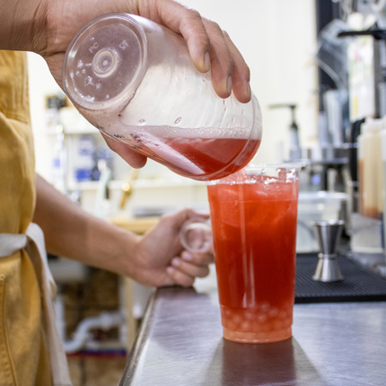 A clear cup with a redish liquid, and lighter colored pearls of boba at the bottom of the glass. Liquid is being poured into the glass from another clear glass  