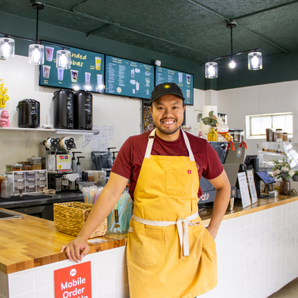 Man smiling at camera, leaning on a counter of a coffee shop. He's wearing a black ball cap, maroon short-sleeved shirt, and yellow apron