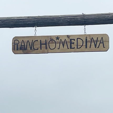 A homemade wooden sign reading "Rancho Medina" in bold, capitalized letters, hanging horizontally from two small chains.