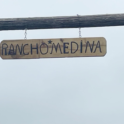 Un letrero de madera hecho en casa que dice "Rancho Medina" en letras mayúsculas y negrita, que cuelga horizontalmente de dos pequeñas cadenas.
