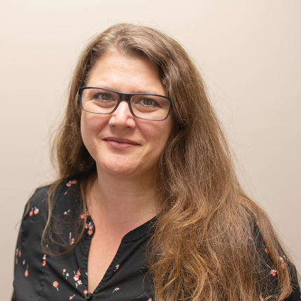 Woman wearing glasses looking at camera with a smile, with long, wavy, brown hair, wearing a black shirt with flowers