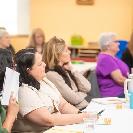 Eight women sit at tables listening to someone speak in front of a room. Items like paper and plastic cups are on the table.