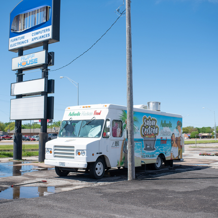A food truck sits between a power pole and a business sign. Food truck has images on the side with one saying "Sabor Costeño"