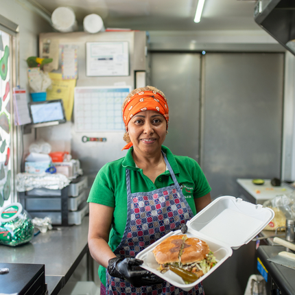 Una mujer latina con un pañuelo rojo, una camisa verde y un delantal a cuadros morado y rosa sostiene un recipiente de poliestireno abierto con un sándwich grande dentro. Ella está parada dentro de un camión de comida con la ventana a la izquierda de la foto.