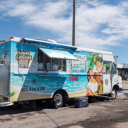 A food truck sits between a power pole and a business sign. Food truck has images on the side with one saying "Sabor Costeño"