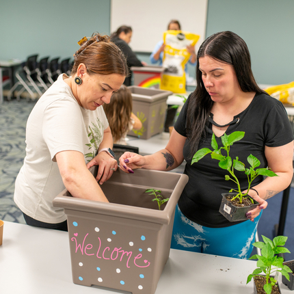 Two Native American women place plants in a square pot with dirt that says "welcome" - the pot is on a table in a carpeted classroom