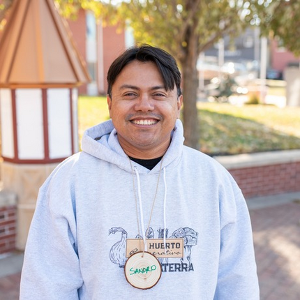 Latino man looking at the camera, wearing a white hooded sweatshirt, a smile, and a wooden name tag that says "Sandro"