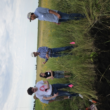 men and women standing in a field with plants up to their wastes, man at center is wearing a blue plaid shirt and white ballcap