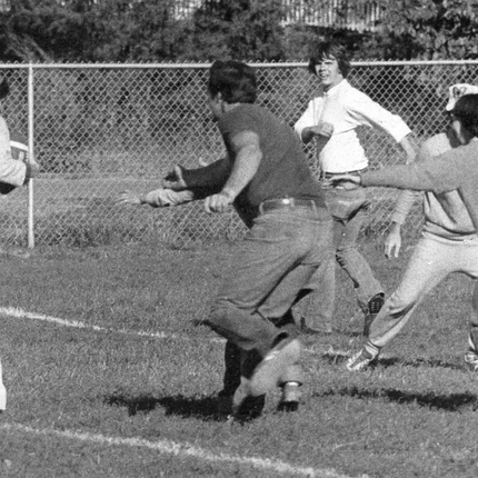Una foto en blanc i negre dels anys 70 d'adults jugant a futbol tàctil. Hi ha 2 dones amb camisa blanca i un home i una dona de gris que es llança cap a la dama de blanc amb la pilota