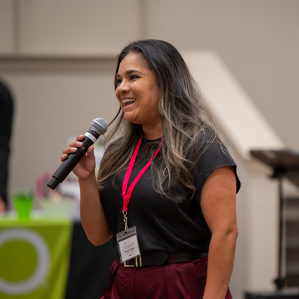 Woman with long brown hair, wearing a black short sleeved shirt, holding a microphone, smiling toward the left of the photo