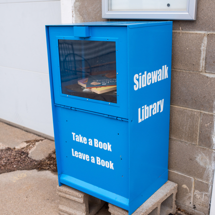 Blue container sitting on top of two cinder blocks in front of a grey brick wall has a window on top half showing a few books inside, white wording on the bottom half and on the side. 