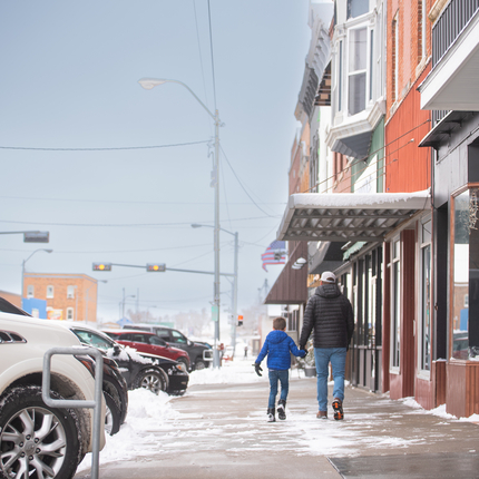 A dad and son hold hands while walking down a small town main street sidewalk. They're wearing coats and snow is swirling on the ground. 