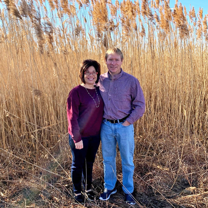 Man and woman standing in front of tall grass in a field. Both are smiling at the camera.