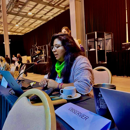 A person with long brownish-black hair, wearing bright green buff and a quarter sleeve button up shirt, speaks into a microphone while seated at a table. In front of them is a computer, a coffee cup, and a name tag that says 'observer' 