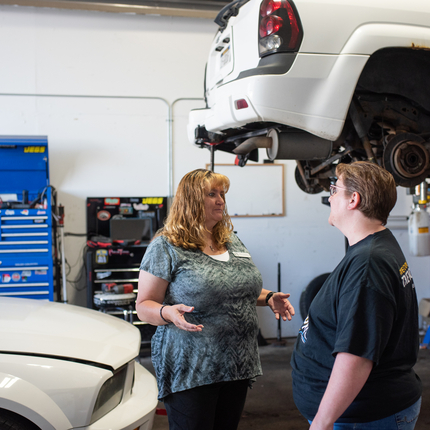 Two women standing in a car repair shop, with one car lifted with its wheels off