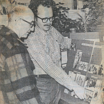पुरानो अखबार फोटो (1970s बाट) दुई पुरुषहरु। प्लेड कोटमा एक जना मानिसले अर्को मानिसलाई प्लेड बटन-अप लामो बाहुलाको शर्टमा फोटोहरू भएको पोस्टरबोर्डमा देखाउँदै हेरिरहेको छ