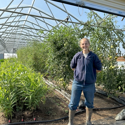 White woman wearing a blue zip up sweater and blue jeans and brown mud boots stands at the entrance of a greenhouse with green plants behind her