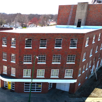 A four-story histrocial brick building with a lot of windows, pictured from above. The building is odd-shaped to fit the corner of an intersection and railroad tracks