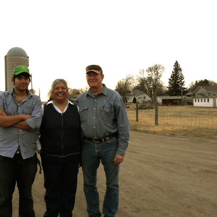 Tres individus: dos homes i una dona, dempeus en una granja davant d'un contenidor de gra amb una casa a la dreta