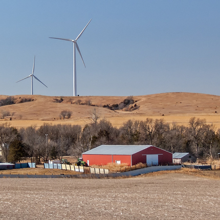 A farm seen across a harvested field, with a red machine shed surrounded by a fence plus a house and several outbuildings. Four wind turbines are behind the farm on a brown hillside.