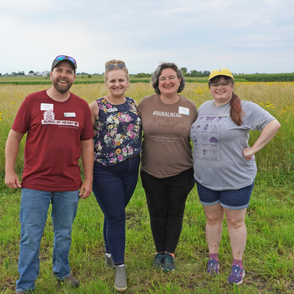 Four individuals standing in a field, a man to the left, and three women.