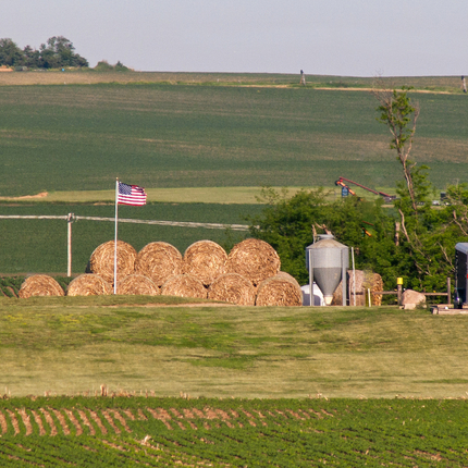 a US flag standing tall over a pile of round hay bales, stacked 2 high. In the foreground and background, there are green fields with short crops.