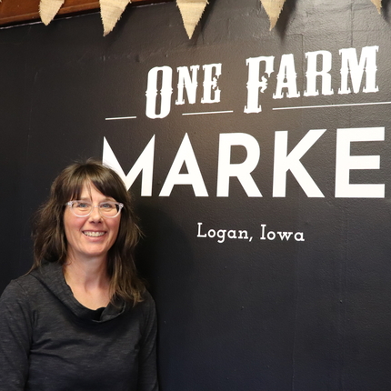 Una dona amb camisa negra amb ulleres transparents es troba davant d'una paret negra amb una escriptura blanca que diu One Farm Market Logan, Iowa
