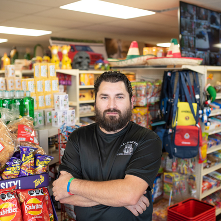 Un home amb barba i camisa negra es troba amb els braços creuats davant de la mercaderia en una botiga