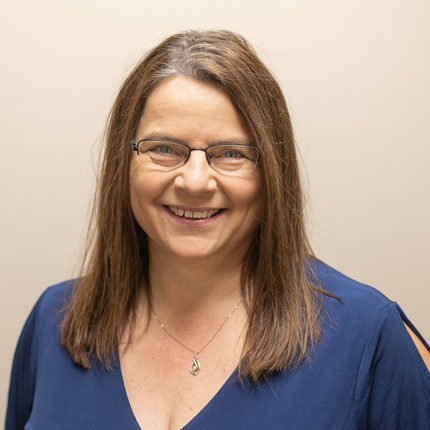 Woman with brown hair and glasses smiling at the camera, wearing a blue shirt and a necklace