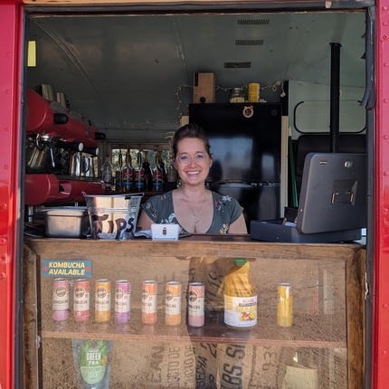 La dona està parada darrere d'un mostrador de begudes de kombucha i una màquina per fer un cafè exprés a la dreta
