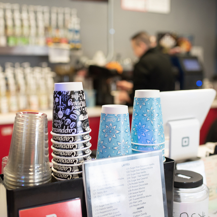 Una fila de tasses, clares, blanques i negres i blaves amb blanc es troben al davant mentre un home treballa una màquina de cafè al fons
