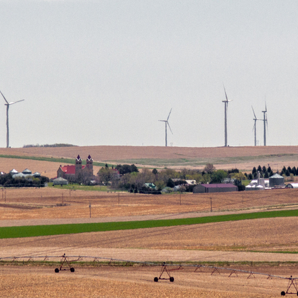 a distant view of a small town in the middle of brown harvested fields, with wind turbines dotting the hills above and behind the town