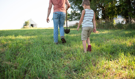A man in jeans, peach shirt, holding a shovel walks through a grass pasture followed by a young boy in tan shorts and a white and tan striped tank top