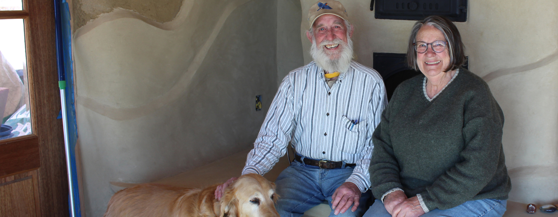 White male with white beard and white female with dark grey hair sit on an indoor bench with a golden dog in front of them