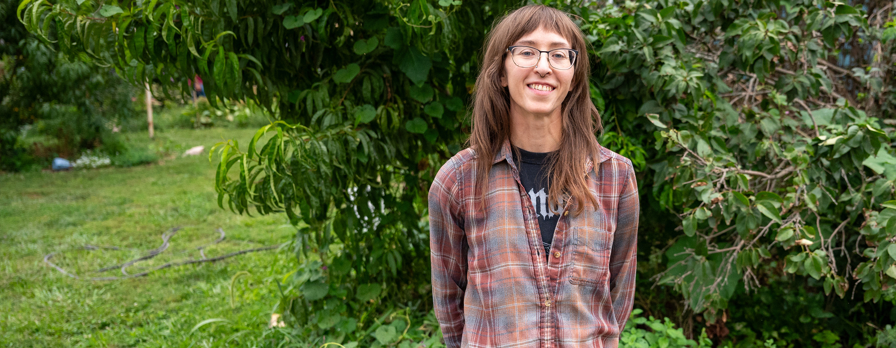 White female in a plaid shirt with long brown hair and glasses stands next to greenery.